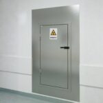 Generator-Lieferantenschleuse von MNT Kwint, wandbündig in Edelstahl 316 eingebaut, geschlossene Tür mit elektronischem ID-Kartenschloss und RADIOAKTIV-Warnzeichen, klinische Umgebung